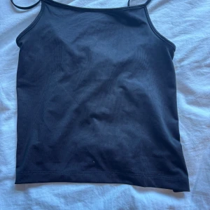 Svart linnetopp med knyt - Säljer en svart linne-topp med smala axelband och har en knytning i ryggen. Toppens minimalistiska design gör den lätt att matcha och den har en tight passform. Perfekt för dig som gillar stilrena och trendiga plagg.