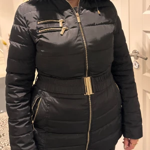 Svart pufferjacka från Rocobarocco - Svart pufferjacka från Rocobarocco med guldfärgade dragkedjor, markerad midja med brett bälte och snyggt foder med leopardmönster. Jackan har flera fickor med dragkedja, huva med fuskpäls och quiltade detaljer. Perfekt för kalla dagar när du vill vara både varm och stylish.
