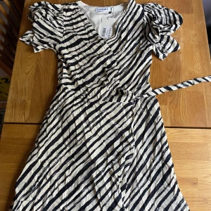 Randig kortklänning från Adoore - Snygg kortklänning från Adoore med svartvita diagonala ränder och puffiga kortärmar. Omlottmodell med knyt i midjan och v-ringad hals. Materialet är mjukt och lätt, perfekt för varma dagar. Klänningen är oanvänd med prislapp.