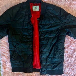 Svart quiltad bomberjacka Jack & Jones - Svart quiltad bomberjacka från Jack & Jones med röd insida. Jackan har dragkedja, ribbade muddar och fickor med knapp. Snygg och enkel modell som passar till det mesta. Perfekt för dig som gillar stilren och sportig look.