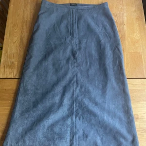 Blå mockakjol från Lindex - Snygg blå långkjol från Lindex i mjuk mocka-look. Kjolen har raka linjer, två bakfickor och en slits bak för extra rörelsefrihet. Perfekt för dig som gillar stilrena och klassiska plagg med en modern touch.