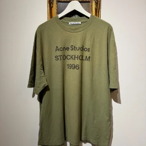 Olivgrön Acne Studios t-shirt L - Olivgrön t-shirt från Acne Studios med svart tryck 'Acne Studios Stockholm 1996' på bröstet. Oversized passform, rund hals och korta ärmar. Tillverkad i mjuk bomull, perfekt för en chill och avslappnad stil.