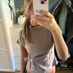 Beige topp med knyt i sidan - Säljer en beige topp med korta ärmar och snygg knytning i sidan. Toppen har en mjuk, stickad känsla och en enkel, avslappnad passform. Perfekt för dig som gillar stilrena och bekväma plagg med en twist.