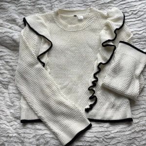 Vit stickad tröja med volang från H&M - Säljer en vit stickad tröja från H&M i storlek S. Tröjan har svarta detaljer längs volangerna på axlarna och vid muddar och nederkant. Eftersom att den är stickad med tjock tråd/snöre håller den än varm på både höst och vintrar, själv har jag använt den till julafton och kalas. Kan upplevas som lite genomskinlig. 