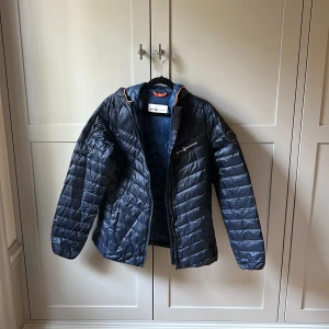 Blå pufferjacka från Sail Racing - Snygg marinblå pufferjacka från Sail Racing med orange detaljer vid huvan. Jackan har quiltad design, dragkedja framtill och logga på bröstet samt ärmen. Perfekt för kyliga dagar och har en sportig vibe. Passar dig som gillar stilren och funktionell ytterjacka. Damjacka men kan nog vara en s/xs för män också