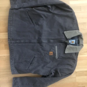 Carhartt brun workwear jacka -  Klassisk brun workwear jacka från Carhartt med kontrasterande beige krage. Jackan har dragkedja framtill, tre fickor och Carhartt-logga på bröstet. Tillverkad i slitstark bomull och har långärmad passform. Perfekt för dig som gillar streetwear och utility-vibe.