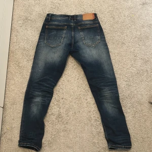 Mörkblå skinny jeans med slitningar - Säljer ett par mörkblåa jeans från Jack & Jones med snygga slitningar och distressed detaljer på benen. Jeansen har klassisk femficksdesign, låg midja och är tillverkade i stretchig bomullsdenim för en bekväm passform.