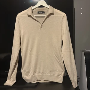 Beige merinoull pikétröja Dressmann - Snygg beige pikétröja från Dressmann i mjuk merinoull. Tröjan har klassisk krage, knäppning framtill och långa ärmar. Perfekt för dig som gillar stilrena och bekväma plagg med en clean look. Otroligt skick 