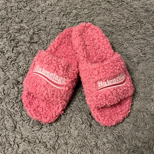 ROSA TOFFLOR  - Mysiga rosa tofflor med broderad logga i vitt, rött och svart på ovandelen. Tillverkade i fluffigt syntetmaterial med platt sula och öppen häl. Perfekta för att chilla hemma med stil.