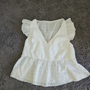 Vit blommig blus med volangärm - Supersöt vit blus med broderade blommor och v-ringning. Toppen har volangärmar och en lätt utsvängd nederdel som ger en luftig känsla. Perfekt för varma dagar och ger en romantisk vibe till din outfit.