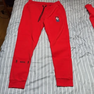 NBA x BOSS mjukisbyxor - Storlek L men passar M! Riktigt snygga röda mjukisbyxor från BOSS i samarbete med NBA. Byxorna har svarta snören i midjan, ribbade muddar och tryck med NBA-logga och BOSS-detaljer på benet. Perfekta för chill eller sportig stil.