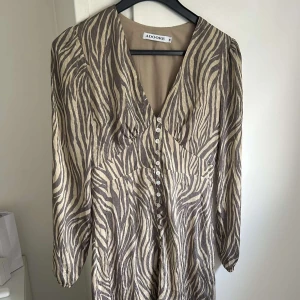 Zebramönstrad klänning från Adoore - Snygg kortklänning från Adoore med zebramönster i beige och grått. Klänningen har v-ringning, knappar framtill🥰