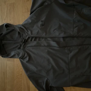 Svart vindjacka från Under Armour - Svart vindjacka från Under Armour med huva och dragkedja framtill. Jackan är tillverkad i lätt syntetmaterial och har en stilren look med diskret logga på bröstet. Perfekt för blåsiga dagar och enkel att matcha med streetwear.