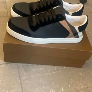 Burberry skor - Snygga svarta sneakers med klassisk rutig detalj i beige, svart och vitt på hälen. Skorna har vita platta sulor, svarta snören och en stilren design som passar till det mesta. Materialmix av skinn och textil för en modern look. Kan byta mot Gucci väska elr keps
