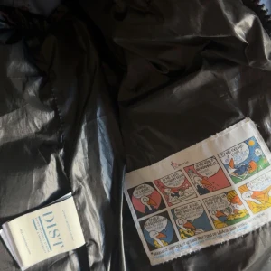 Svart dunväst från Moncler - Snygg svart dunväst från Moncler med glansig finish och klassisk logga på bröstet. Västen har två fickor med dragkedja och en cool serietidningsdetalj på insidan. Perfekt för lager-på-lager och streetstyle. Fylld med vitt gåsdun för extra värme. Och har NFC tag 