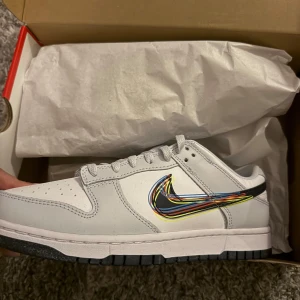 Nike Dunk Low med färgglad swoosh - Nike Dunk Low sneakers i vitt och ljusgrått skinn med svart sula. Swoosh-loggan är unik med flera färgglada linjer i blått, gult, rött och orange. Klassisk siluett med snörning och rund tå. 
