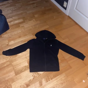 Ralph lauren hoodie - Svart hoodie från Polo Ralph Lauren med dragkedja framtill och klassisk huva. Tröjan har långa ärmar och den ikoniska Polo-loggan broderad på bröstet. Perfekt för chill dagar eller när du vill ha en enkel och stilren look.