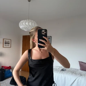 Svart satin linnetopp från Gina Tricot - Svart linnetopp i glansigt satinmaterial från Gina Tricot. Toppen har breda axelband, fyrkantig halsringning och snygg snörning i sidan för att justera passformen. Perfekt att styla med jeans eller kjol för en trendig look.