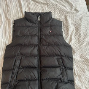 Svart dunväst från GANT - Svart dunväst från GANT med hög krage och diskret logga på bröstet. Västen har två fickor framtill och stängs med dragkedja. Den quiltade designen ger en snygg och sportig look, perfekt att slänga över en hoodie när det är kyligt ute. Köpt för 1200 kr 