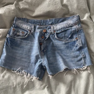 Blå jeansshorts från Vance Vintage - Säljer ett par blå jeansshorts från Vance Vintage med fransig och rå kant nertill. Shortsen har klassisk femficksdesign, knappar framtill och coola slitningar för en avslappnad vibe. Tillverkade i 100% bomull,