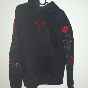 Svart hoodie med rosbroderi från H&M - Svart hoodie från H&M Divided i storlek XS med broderade röda rosor och gröna blad längs båda ärmarna. Framsidan har texten 'LOVE IS BLIND' i rött. Tröjan har huva med snörning och en stor magficka. Perfekt för dig som gillar detaljer och blommigt.