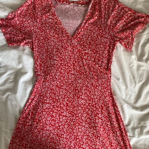 Röd blommig omlottklänning Indiska XS - Supersöt röd kortklänning från Indiska med vita småblommor, omlottmodell och v-ringning. Klänningen har korta ärmar och markerad midja, perfekt för sommarens alla tillfällen. Lätt och skön i materialet.