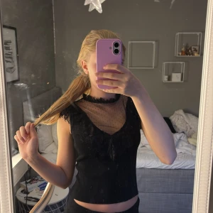 Svart meshblus med spets och prickar - Supersnygg svart blus med transparent mesh upptill och små prickar. Axelpartiet och halsringningen har volangdetaljer och spets, vilket ger en cool och romantisk vibe. Finns en knapp i nacken som får den att sitta lite bättre framtill. Ärmlös och croppad modell som passar perfekt till höga jeans eller kjol.