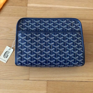 Goy4rd Handväska - Säljer en blå handväska med deras ikoniska grafiska mönster i vitt och grått. Väskan har silverfärgad dragkedja, vita kontrastsömmar och detaljer i skinn. Perfekt för att bära dina accessoarer med stil. 