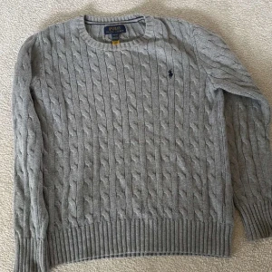 Grå kabelstickad tröja Polo Ralph Lauren - Kabelstickad grå tröja från Polo Ralph Lauren med klassisk rund hals och broderad logga på bröstet. Långärmad modell med ribbade muddar och nederkant. Perfekt för dig som gillar en stilren och tidlös look.