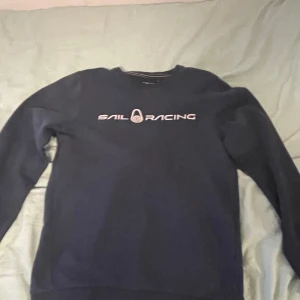 Mörkblå Sail Racing sweatshirt - Mörkblå sweatshirt från Sail Racing med vit logga tryckt på bröstet och en mindre logga i nacken. Klassisk rund hals och långa ärmar, perfekt för en chill och sportig look. Skön passform och stilren design som funkar året runt.
