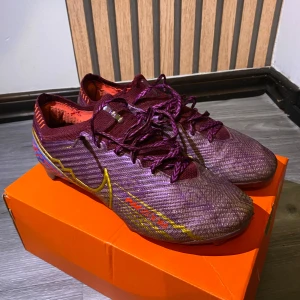 Vinröda Nike fotbollsskor Zoom Mercurial - Snygga vinröda Nike elite fotbollsskor Zoom Mercurial med guldiga detaljer och lila mönster. Skorna har snörning, låg profil och är tillverkade i syntetmaterial för bästa grepp och känsla på planen. Perfekta för dig som vill sticka ut med stil på matchen. Pris kan diskuteras!