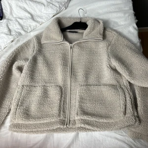 Beige teddyjacka från 157, storlek S - Mysig beige teddyjacka från 157 i storlek S. Jackan har dragkedja framtill, två stora fickor och hög krage. Perfekt för kyliga dagar när du vill vara både varm och stilren. Fleece-liknande material som känns supermjukt.