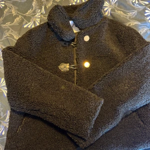 Svart teddyjacka från Zara, S - Mysig svart teddyjacka från Zara i storlek S. Jackan har stora knappar och snygga detaljer i skinnimitation vid knäppningen och fickorna. Perfekt för kalla dagar med sin fluffiga och trendiga look. Hög krage och långärmad modell.