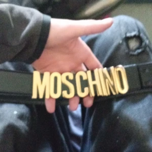 Svart Moschino bälte i läder - Säljer ett svart bälte från Moschino i äkta läder med stora, guldiga bokstäver framtill. Bältet är stilrent och har en bred design som verkligen sticker ut. Perfekt för dig som vill ha något lite mer annorlunda men fortfarande stilrent. Bältet är äkta och köpt på zalando för 3000kr