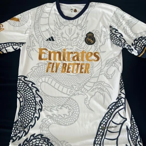Real Madrid Ronaldo draktröja - Snygg Real Madrid fotbollströja med Ronaldo 7 på ryggen. Vit bas med grått och svart drakmönster, guldiga detaljer och tryck. Kortärmad med mörkblå krage och Aeroready-material som andas. Perfekt för dig som vill sticka ut på planen eller läktaren. 