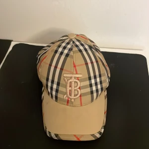 Burberry rutig keps med logga - Snygg keps från Burberry med klassiskt rutigt mönster i beige, svart, vitt och röda detaljer. Framtill sitter en broderad TB-logga i beige. Justerbar rem baktill och svart insida med Burberry-tryck. Perfekt för dig som gillar lyxiga accessoarer.