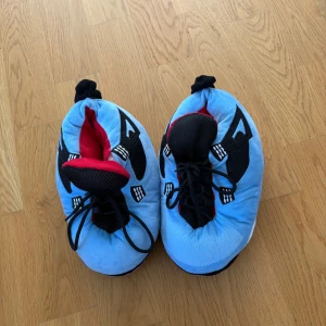 Blå fluffiga sneakers-tofflor Jordan 44  - Supermjuka och oversized sneakers-inspirerade tofflor i ljusblått med svarta och röda detaljer. Insidan är röd och ovandelen har snörning och mesh-liknande tyg. Sulan är svart med små gummiprickar för extra grepp. Perfekta för att chilla hemma med stil. Är liknande Jordan innetofflor och de är köpta för 579kr på SCORETT men mitt pris är 149kr