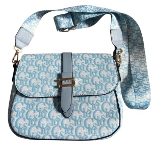Blå axelväska  - Snygg axelväska  i ljusblått och vitt med tydligt monogrammönster. Väskan har bred justerbar axelrem, guldfärgade detaljer och flera fack med dragkedja. Materialet är en mix av syntet och textil, perfekt för dig som gillar statement-accessoarer.