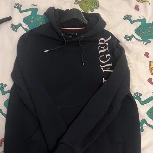 Marinblå hoodie Tommy Hilfiger M - Snygg marinblå hoodie från Tommy Hilfiger i storlek M. Tröjan har stor vit logga längs ena sidan framtill, huva med snörning och är tillverkad i en mjuk blandning av bomull och polyester. Perfekt för chill dagar eller när du vill ha en clean streetstyle.