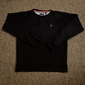 Mörkblå sweatshirt från GANT - Mörkblå sweatshirt från GANT med klassisk rund hals och broderad logga på bröstet. Tröjan har långa ärmar, ribbade muddar och nederkant samt en stilren look. Perfekt för dig som gillar enkel och tidlös stil. Storlek 170. Hör av er vid frågor och funderingar😄🙌