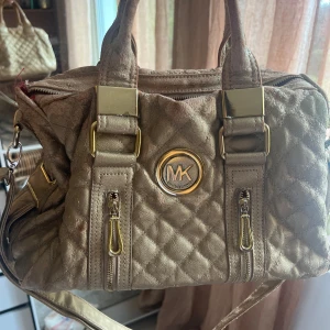 Quiltad guld handväska Michael Kors - Säljer en snygg quiltad handväska från Michael Kors i guldigt skinn med metallic finish. Väskan har två handtag, axelrem, guldfärgade detaljer och MK-logga framtill. Utsidan har två dragkedjor och insidan är fodrad i rött. Perfekt för dig som gillar lyxig stil.