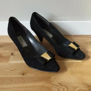 Svarta mockapumps från Gabor - Snygga svarta pumps från Gabor i mjuk mocka med spetsig tå och låg klack. Dekorerade med en svart rosett och guldfärgad detalj framtill. Behöver klackas om, klacl ca 7cm. Fler bilder på Vinted