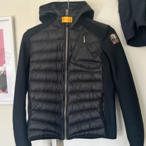 Svart pufferjacka från Parajumpers - Svart pufferjacka från Parajumpers med huva och coolt badge på axeln. Jackan har quiltad framsida och rygg, stickade ärmar och flera dragkedjeförsedda fickor. Snygga detaljer som gul rem vid halsen och ribbad struktur på ärmarna.