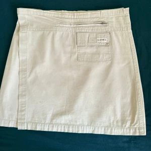 Beige omlottkjol - Stilren beige omlottkjol från Camel i bomull. Kjolen har praktisk ficka med lock och dragkedja baktill samt kardborrestängning i midjan. Perfekt för dig som gillar enkel och clean stil med funktionella detaljer. Kjolen passar i M.