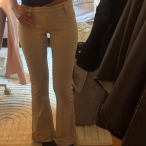 Beiga bootcut byxor med låg midja - Snygga beiga kostym byxor med bootcut och låg midja. Byxorna har två knappar framtill och sitter tajt över låren innan de släpper ut vid bensluten. 