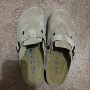 Beige mocka Birkenstock sandaler - Säljer ett par klassiska Birkenstock sandaler i beige mocka med justerbart spänne och platt sula. Skorna har en rund tå och svart räfflad yttersula för extra grepp. Perfekta för chill dagar och enkel stil. Sista bilden är med ljus