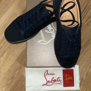 Svarta sneakers från Christian Louboutin - Snygga svarta sneakers från Christian Louboutin i mocka med klassisk röd sula. Skorna har rund tå, svarta skosnören och diskret broderad logga på hälen. Lyxig känsla och stilren design som sticker ut med den ikoniska röda undersidan.
