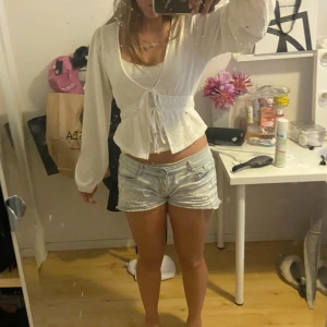 Vit omlottopp med knyt och ballongärm - Supersöt vit omlottopp med v-ringning och knyt i midjan. Toppen har långa ballongärmar och är lätt croppad med en smickrande passform. Materialet är strukturerat och känns luftigt, perfekt för varmare dagar. Enkel att styla med både jeans och kjol.