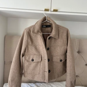 Beige ulljacka från Zara - Snygg beige jacka från Zara i ullblandning med klassisk krage och stora knappar framtill. Jackan har två stora fickor med lock och en croppad passform som ger en trendig look. Perfekt för lager-på-lager-stil under höst och vinter.passar mig som annars bär S perfekt.