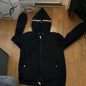 Svart vindjacka från Moncler med  helt ny - Svart vindjacka från Moncler med huva och dragkedja. Jackan har Moncler-logga på ärmen och snygga röd, vit och blå ränder på huvan. Tillverkad i glansig polyester, perfekt för blåsiga dagar. Fickor med tryckknappar och stilren look. 